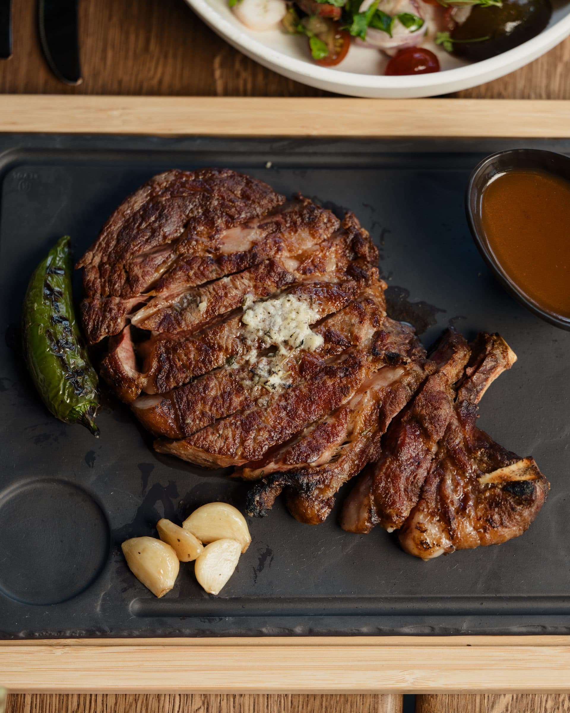 Dry-aged ribeye on a slate board with roasted garlic and a grilled pepper